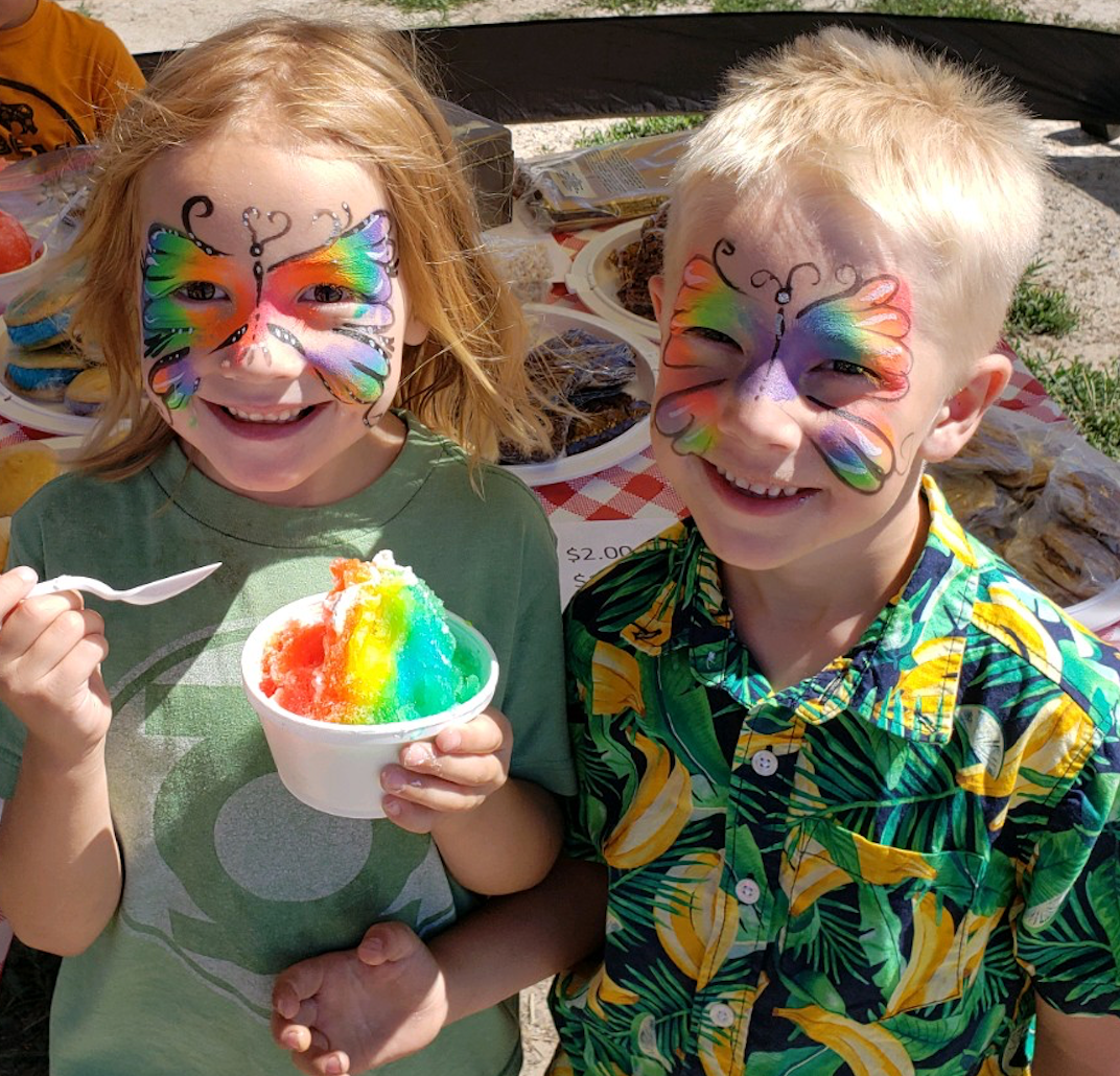 Two children with butterfly face paint sharing a rainbow snow cone at the market